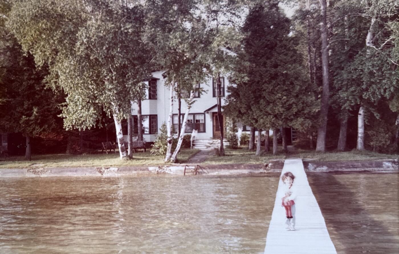 A toddler standing on a dock with a red doll in her arms. Behind her, a house with green shutters and birch trees in the front yard.