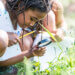A multi-ethnic group of elementary school children are outdoors on a sunny day. They are wearing casual clothing. They are learning about nature in science class. A girl of African descent is using a magnifying glass to look for bugs.
