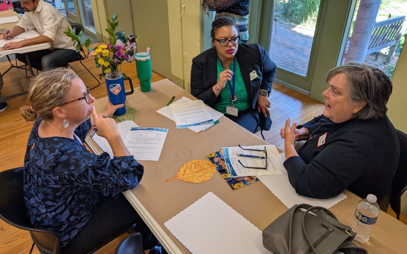 Three women sit around a table discussing papers, with flowers and water bottles nearby.