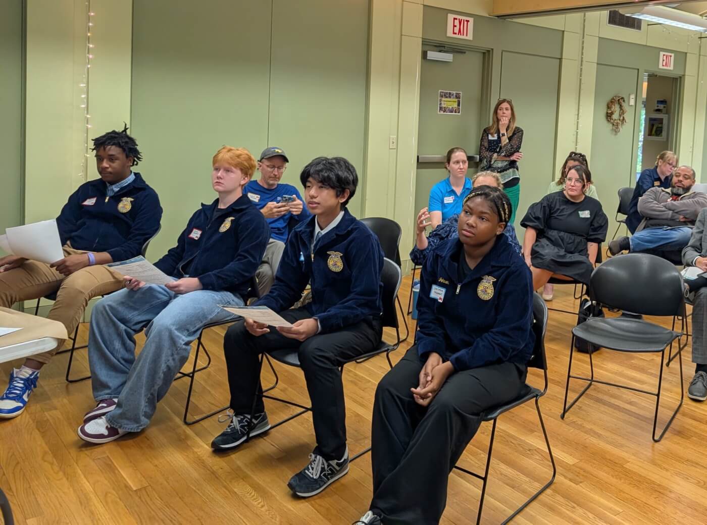A group of students sit in chairs holding papers while adults watch from the back of the room.