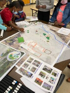 Five students lean over a map of their schoolyard on a table. They place color-coded stickers on certain areas of the map. 