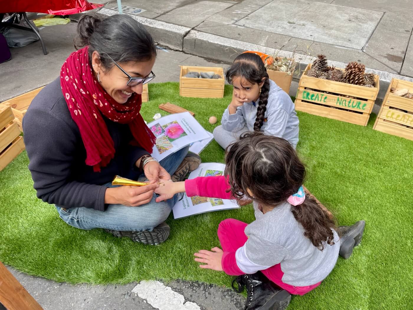A woman paints a child’s hand while two kids watch during an outdoor nature activity.