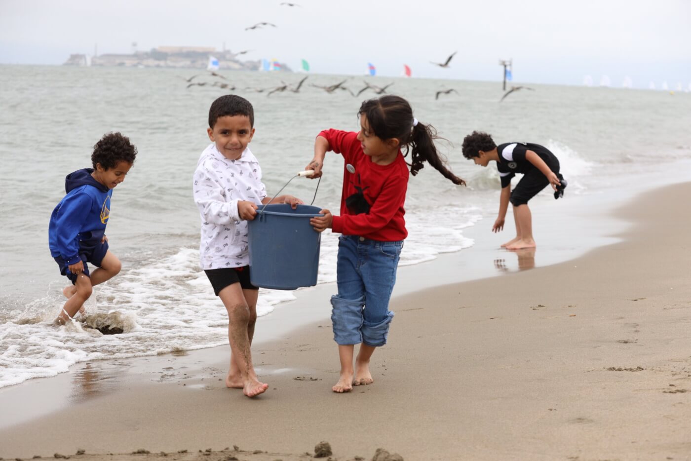 Kids play at the beach, carrying a bucket and splashing in the waves.