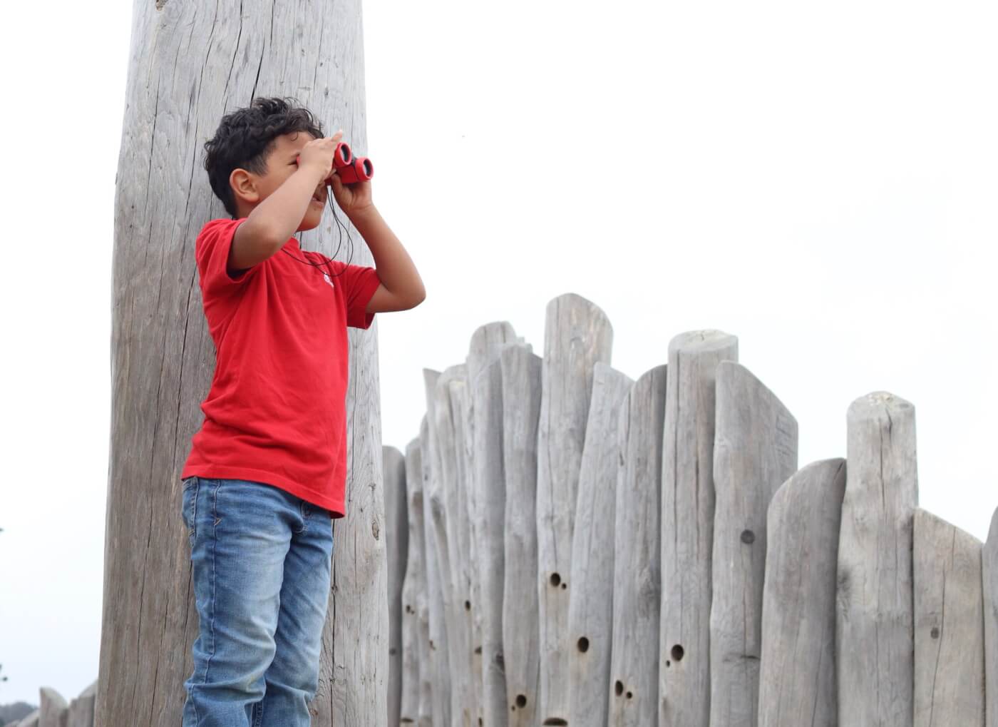 A child uses binoculars while standing by a wooden fence.