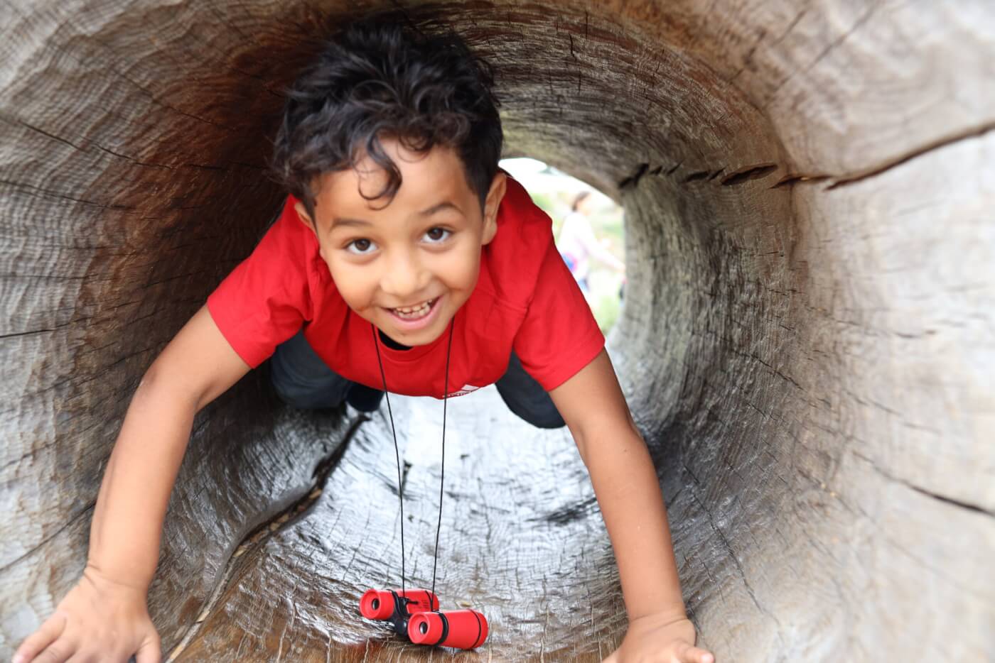 A child crawls through a hollow log, smiling at the camera.