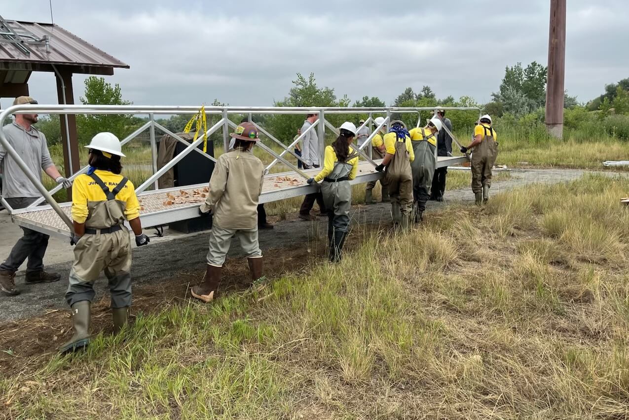 A group of workers in helmets and waders carry a large metal structure across a grassy area.