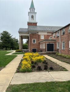 Brick church building with white steeple, columns, and yellow flower beds along walkway.