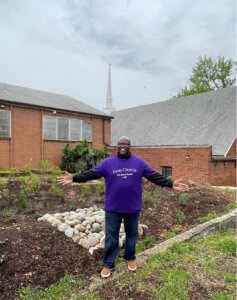 Man standing with arms outstretched in front of a church rain garden and native landscaping.