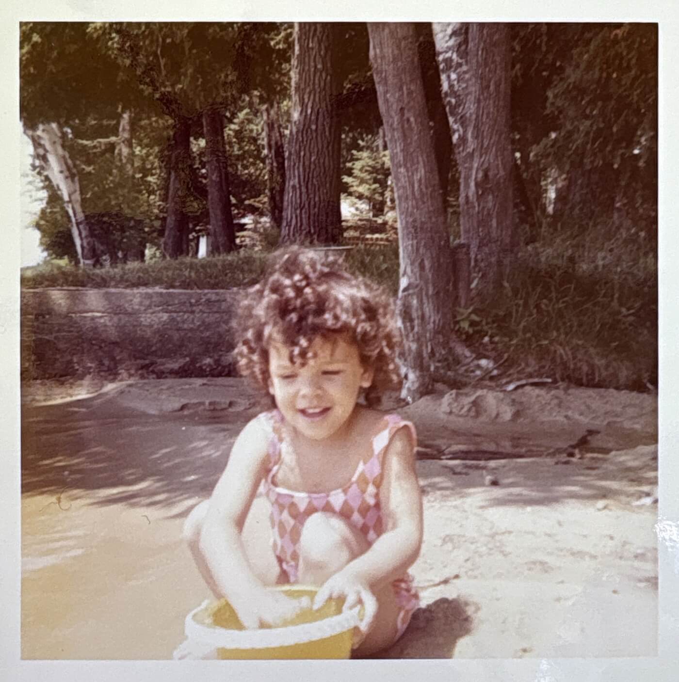 A toddler with curly black hair and a pink checkered bathing suit plays with a yellow bucket of sand.