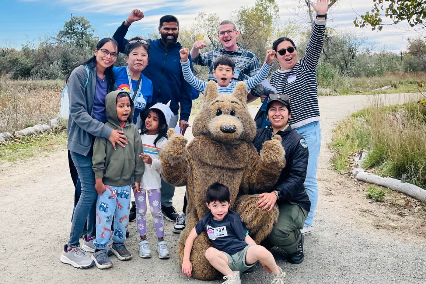 A group of adults and children pose outdoors on a trail with a person in a bear costume, smiling and raising their arms.