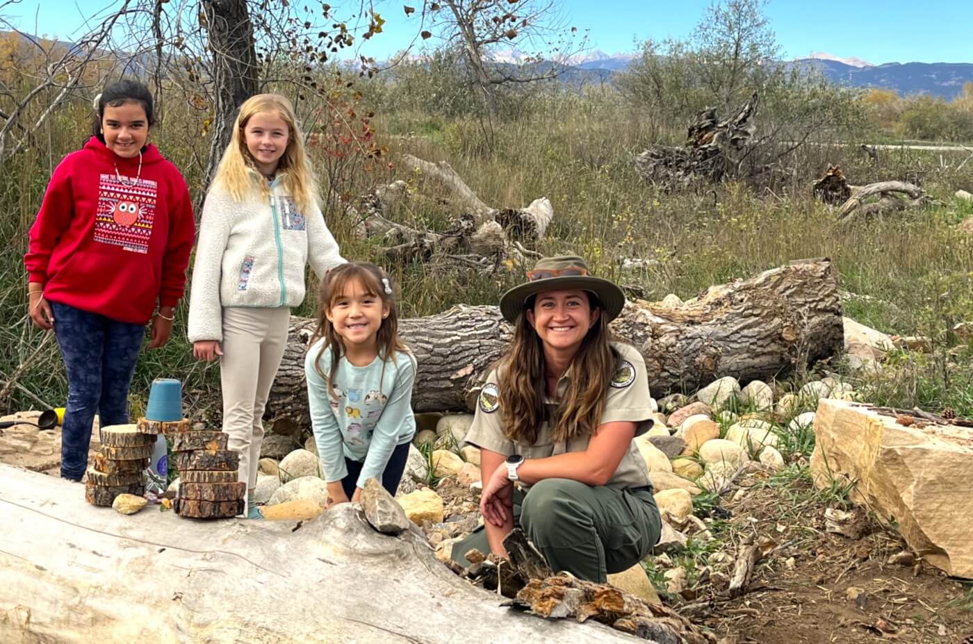 Three children and a park ranger pose outdoors near logs and rocks with mountains in the background.