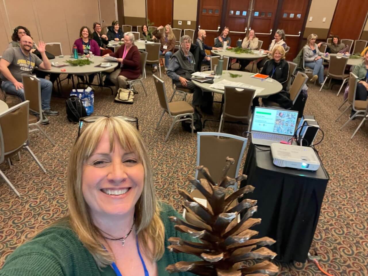 Maureen takes a selfie with a pinecone in front of a conference room, where an audience is seated at several circular tables.