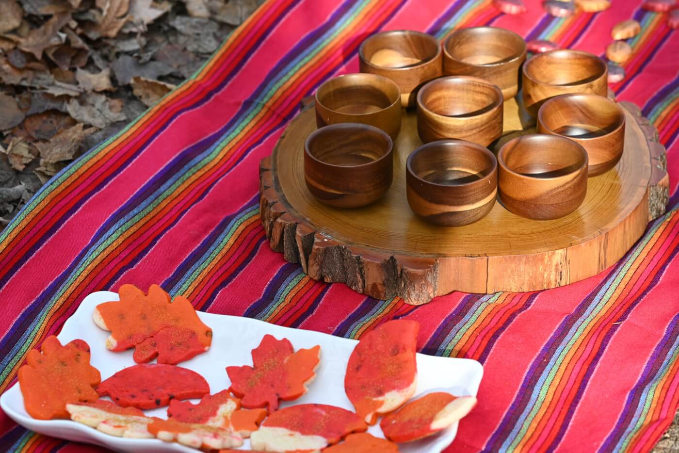 A white plate with orange leaf-shaped cookies sits on a colorful table cloth, next to a tree round serving platter with nine wooden cups.