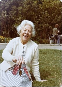 A vintage photograph of an older woman with curled hair and a white cardigan, sitting on a lawn.