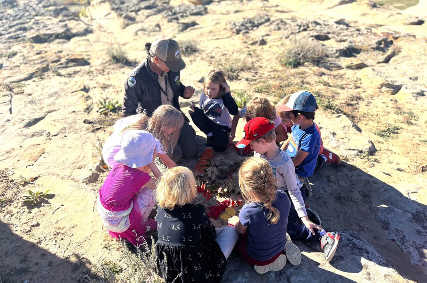 A ranger kneels with a group of young children gathered in a circle on rocky ground, looking at leaves and natural objects.