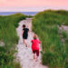 Two young boys running together down a sandy path between tall grass near a beach head.