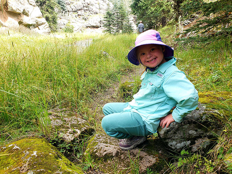 A young child wearing a light blue shirt, matching pants, and a sun hat crouches on a grassy trail in a forested area. The child is smiling brightly at the camera. The background features tall grass, evergreen trees, and rocky cliffs.