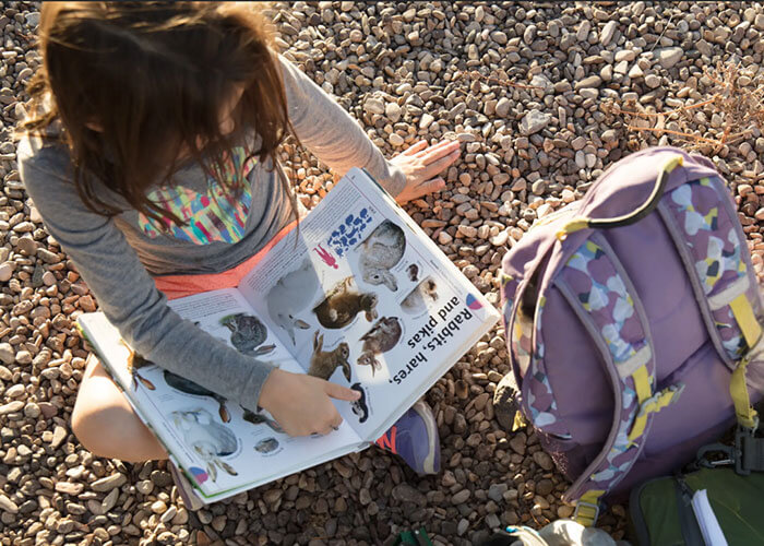 Young child reading a book next to her backpack while sitting on rocks.
