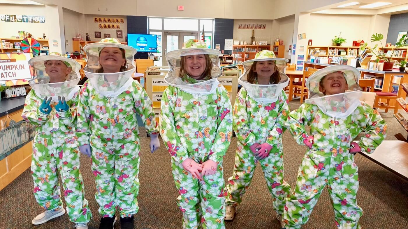 Five students pose inside a school library, donned in colorful green and pink beekeeping suits.