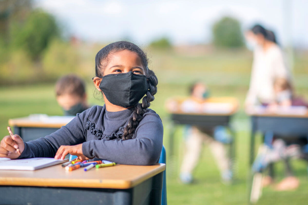 Young girl with mask sitting at a desk outside.