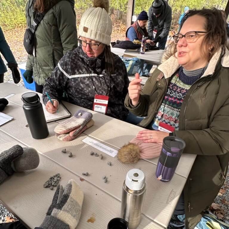 Two teachers sit at a picnic bench. One writes on in a notebook with a pencil, the other is in conversation. On the table are several different piles of small rocks.