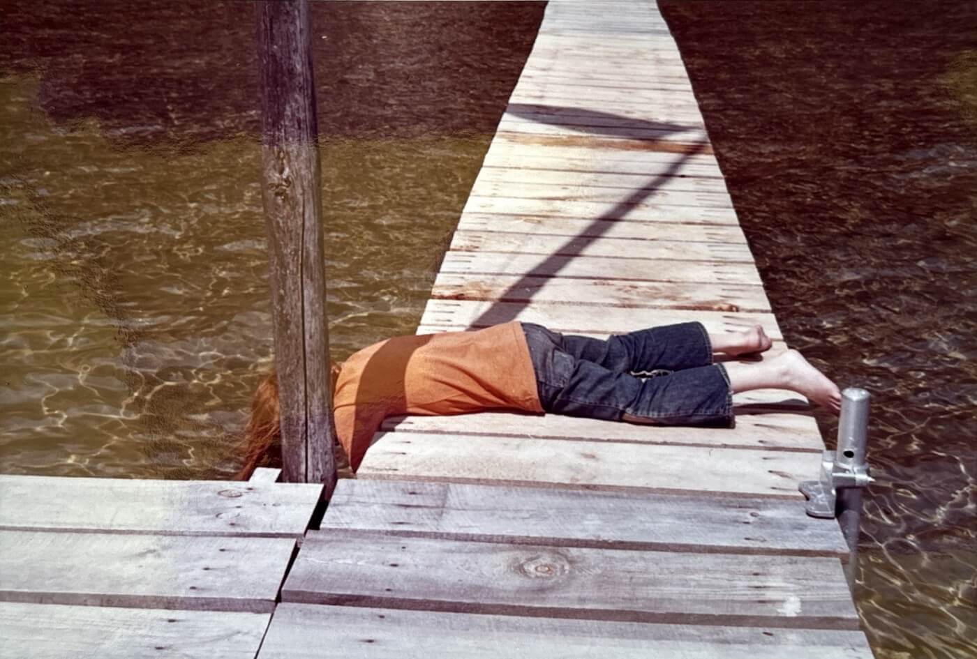 A boy with red hair lays across the walkway of a deck, looking under it.