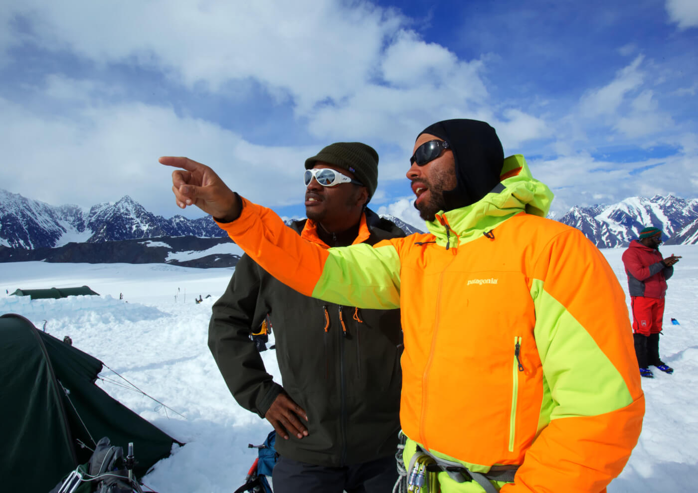 Two African American men, dressed in warm clothing in the snowy mountains with tents next to them. One man is pointing to the distance.