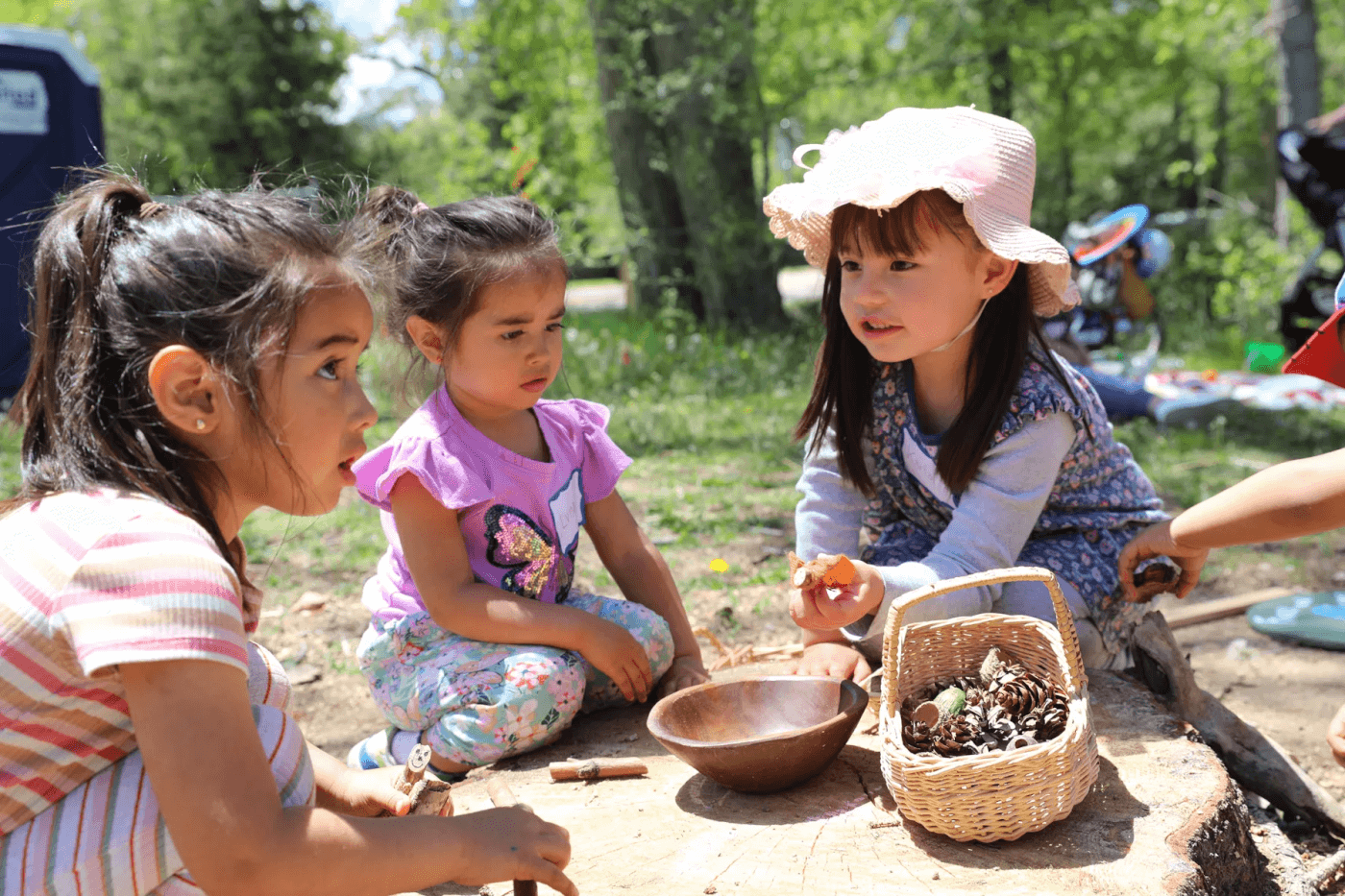Three young children sit outdoors on a tree stump, playing with pinecones and small objects from a basket.