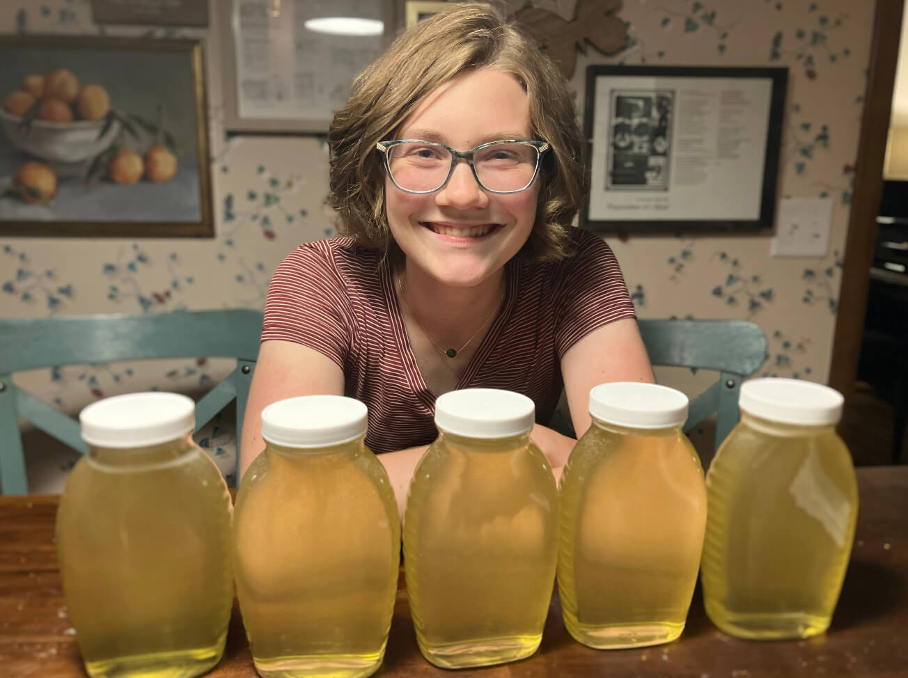 Audrey smiles in her home, seated behind five glass containers of pale yellow honey.