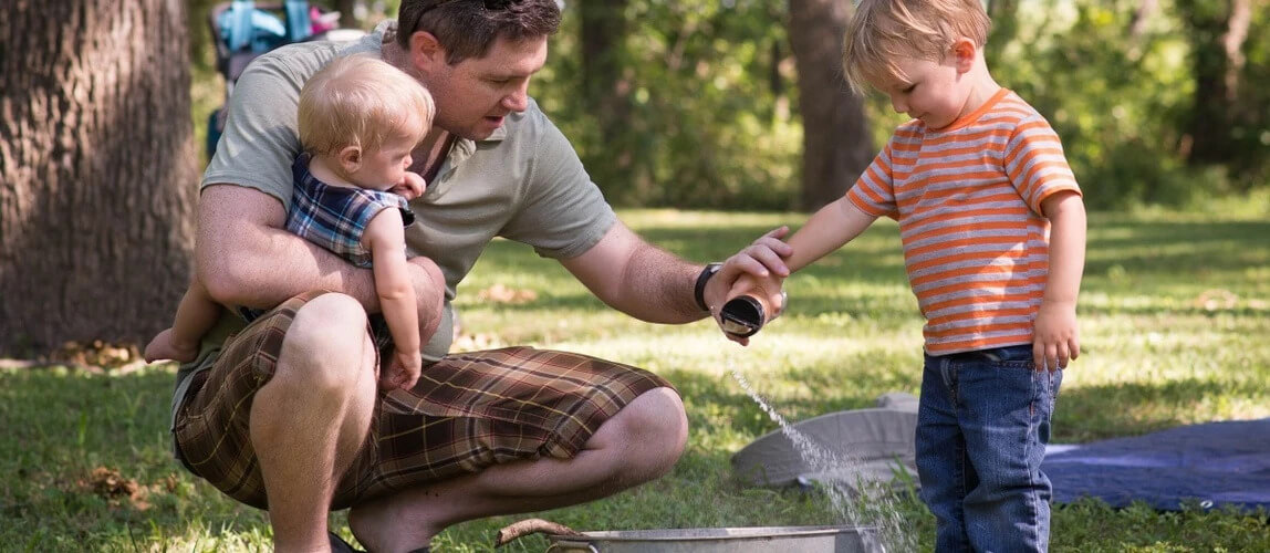 father holding one child and helping another pour water in a bucket