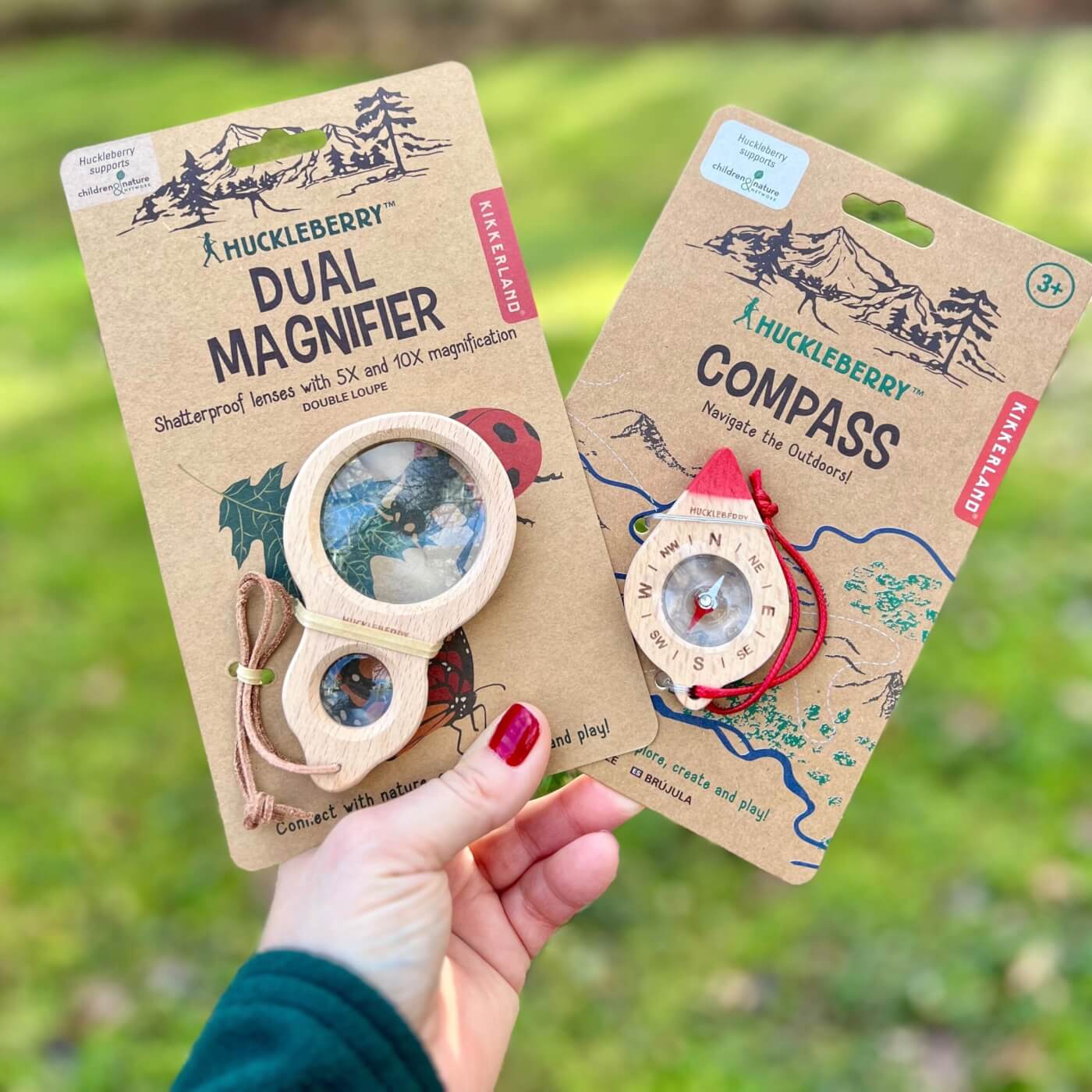 two huckleberry products including a dual magnifier and a compass, held in persons hand with nature in the background.