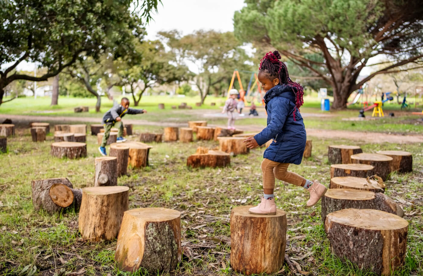 Child stepping across tree stumps in a park while other children play in the background.