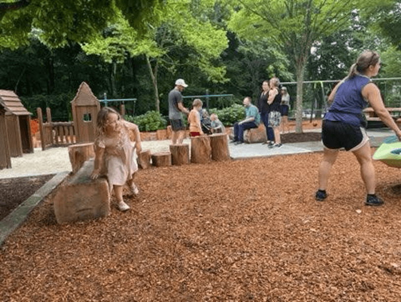 Children and families play and gather at Peterson Park’s natural playground with wooden structures and shaded trees. Photo credit: Providence Parks Department.