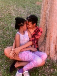 A mother and her daughter cuddle by a tree.