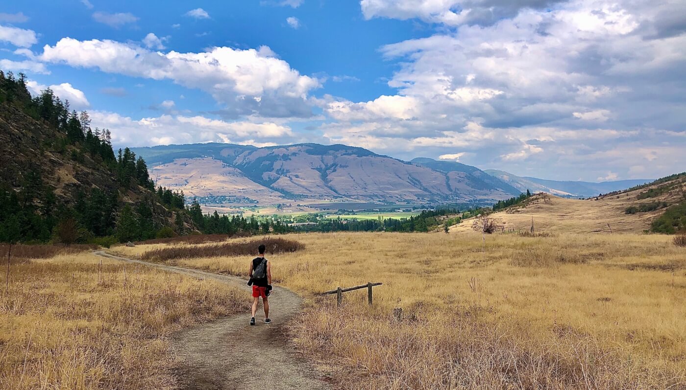 A person walks on a winding dirt trail through a dry, grassy field leading towards distant mountains.