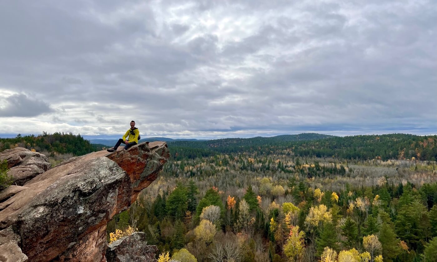 An individual in a yellow jacket sits on the edge of a reddish rock cliff above a forest with changing leaves and rolling hills.