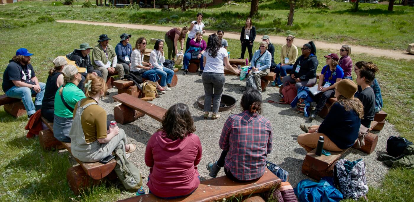 large group of people sitting in a circle surrounding a woman who is standing and speaking to the group