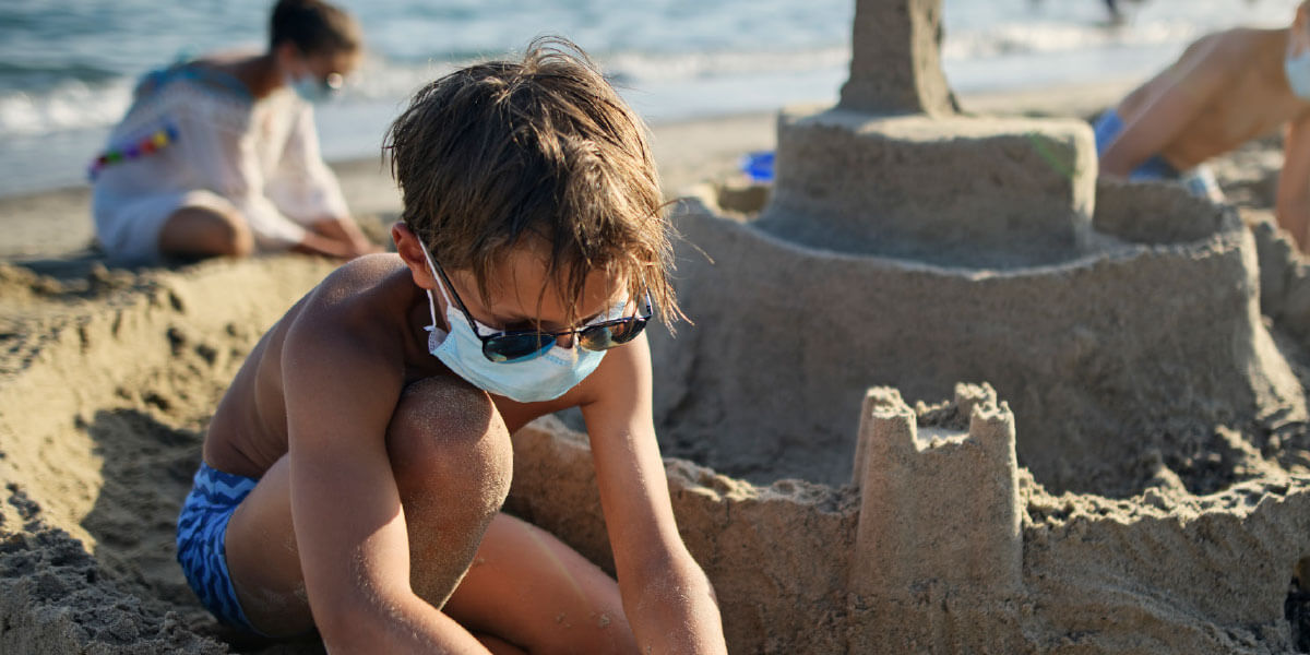 Boy playing in the sand, making a sand castle while wearing a mask.