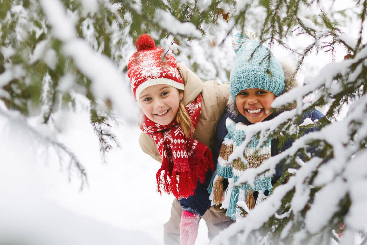 Two kids smiling outside in the behind a snowy tree branch during the winter.