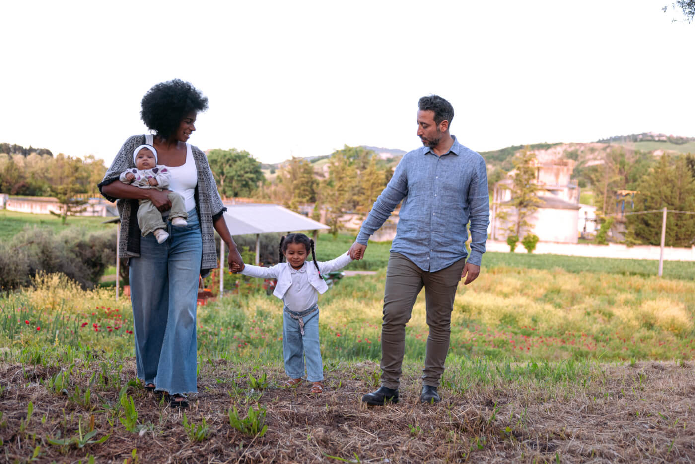 Family of four walking together through a sunlit meadow at dusk, parents holding hands with a young daughter while the mother carries a baby.Family of four walking together through a sunlit meadow at dusk, parents holding hands with a young daughter while the mother carries a baby.