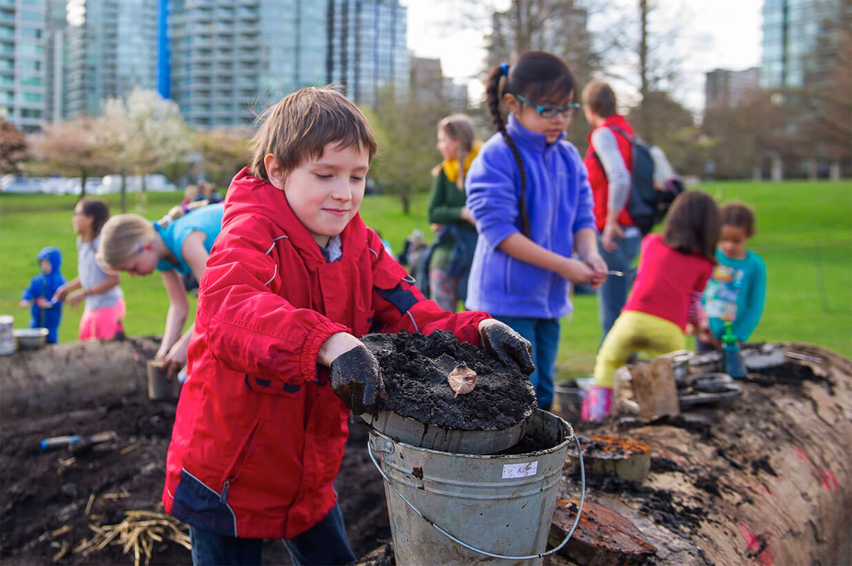 Young boy with bucket full of dirt digging with other children.