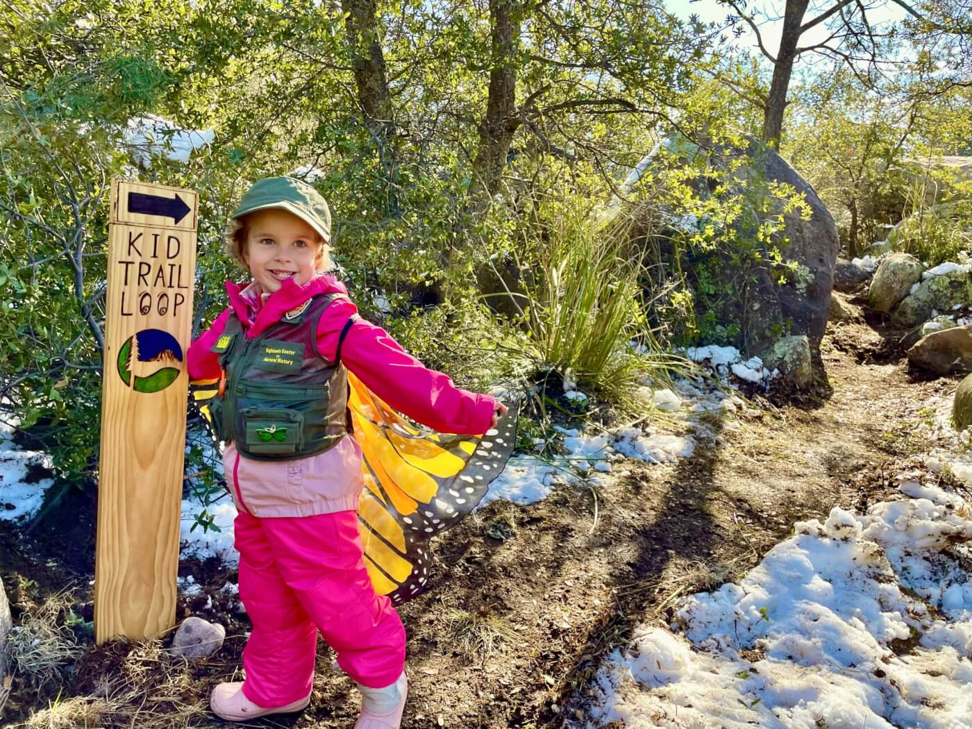 little girl smiling at the camera, with a cape on that is similar to monarch butterfly wings. She is standing next to a sign that says "kid trail loop"