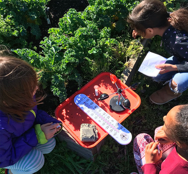 Three young children examining a garden with a thermometer, notepads, and other tools.