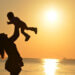Woman carrying baby at beach during sunset.